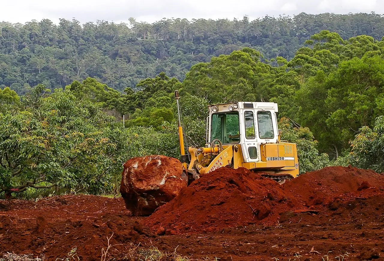 Bulldozer pushing a large boulder through red soil beside trees